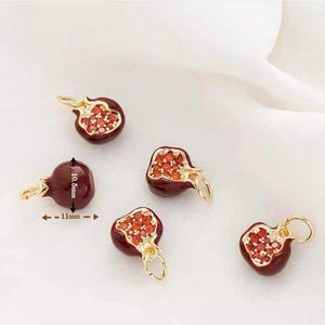 May include: Five pomegranate-shaped charms. Each charm is a deep red color with a gold-colored bail. The pomegranate charms are approximately 0.4 inches wide and 0.4 inches tall. The inside of the pomegranate is filled with small, red, gem-like stones.