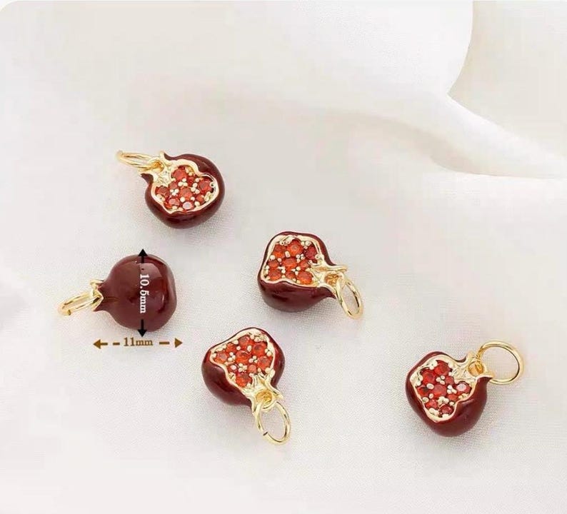 May include: Five pomegranate-shaped charms. Each charm is a deep red color with a gold-colored bail. The pomegranate charms are approximately 0.4 inches wide and 0.4 inches tall. The inside of the pomegranate is filled with small, red, gem-like stones.