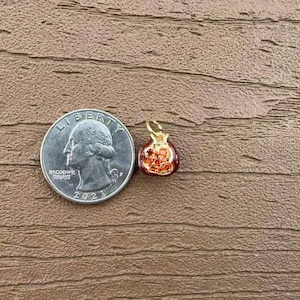 May include: A small, red and gold pomegranate charm with a gold loop, next to a US quarter. The charm has a detailed design with red and orange accents, and is set against a brown wooden background. The quarter is dated 2021.
