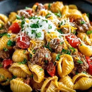 May include: A close-up shot of a pasta dish with shell-shaped pasta, ground meat, diced tomatoes, shredded cheese, sour cream, and chopped parsley. The dish is served in a dark bowl, with a variety of textures and colors.