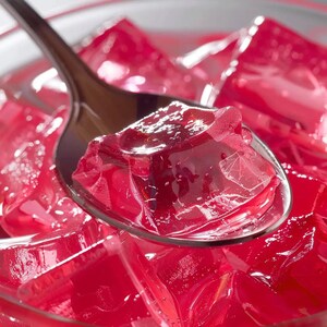 May include: Close-up of a spoonful of red gelatin dessert, with a clear glass bowl in the background. The gelatin is cut into cubes and has a glossy, transparent appearance. The spoon is dark brown.