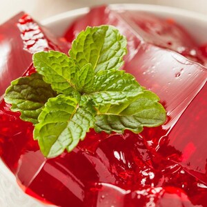 May include: A close-up shot of a white bowl filled with red gelatin cubes, garnished with fresh green mint leaves. The gelatin has a glossy, transparent appearance. The dessert is a vibrant red color, contrasting with the white bowl and green mint.