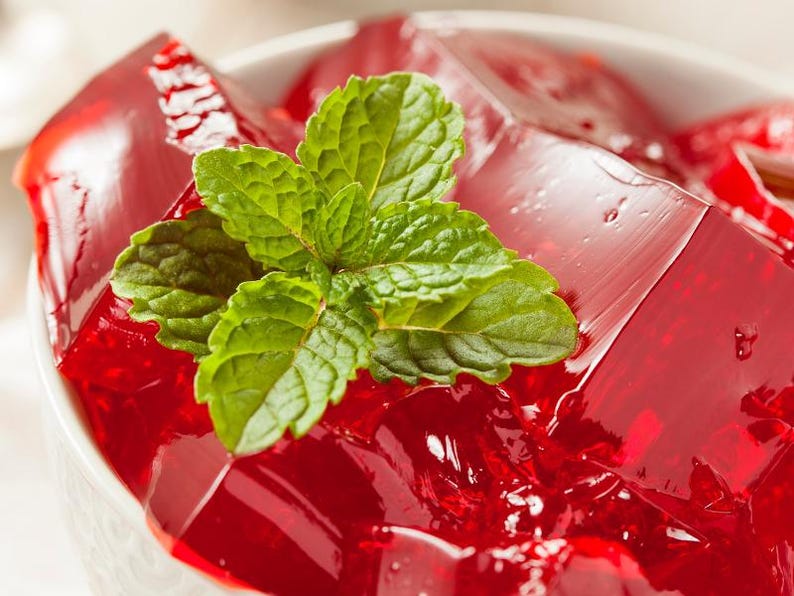 May include: A close-up shot of a white bowl filled with red gelatin cubes, garnished with fresh green mint leaves. The gelatin has a glossy, transparent appearance. The dessert is a vibrant red color, contrasting with the white bowl and green mint.
