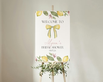 May include: A white sign on a wooden easel with the text "WELCOME TO Alyssa's BRIDAL SHOWER 01.05.26". The sign is decorated with watercolor lemons, leaves, and a yellow bow.