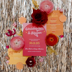 Op de afbeelding: Een trouwuitnodigingssuite met een roze en oranje kleurenschema. De uitnodigingskaart leest "Christopher & Megan" met de datum "10.11.25". De suite bevat een ringdoos, menu en decoratieve bloemen tegen een zilveren slingerafstand.