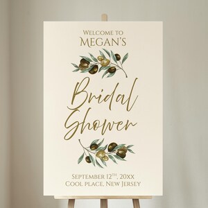 May include: A cream-colored welcome sign on a wooden easel. The sign reads "Welcome to Megan's Bridal Shower" in elegant gold script, adorned with olive branch illustrations. The date and location are also displayed.