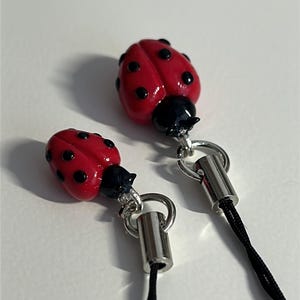 May include: Two ladybug-shaped charms with red bodies and black spots. Each charm has a black head and is attached to a silver-colored metal loop and a black cord. The charms are set against a white background.