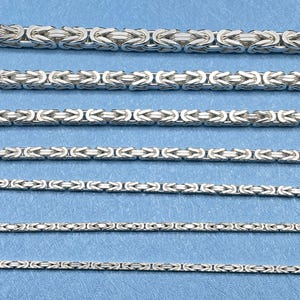 May include: Close-up of several silver Byzantine chain necklaces of varying thicknesses. The chains are displayed against a light blue background. The links are intricately woven, creating a textured appearance. The chains are likely made of sterling silver.