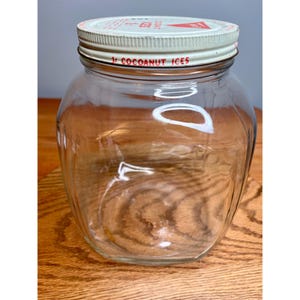 May include: A clear glass jar with a metal lid. The lid is white with red and blue text that reads "1¢ COCOANUT ICES". The jar is empty and sits on a wooden surface.
