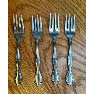 May include: A set of four identical silver-toned metal forks arranged in a row. Each fork features a three-tined head and a slender handle that widens toward the base, showcasing a smooth, polished finish with subtle decorative curves.