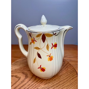 May include: Cream-colored ceramic pitcher with a lid and a curved handle. The pitcher is decorated with a floral pattern in shades of red, yellow, and brown. The rim and handle are trimmed with gold. The pitcher is sitting on a wooden surface.
