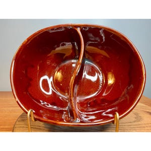 May include: A divided serving bowl with a rich, glossy, reddish-brown glaze. The oval-shaped bowl is separated into two compartments by a central divider. The bowl is displayed on a gold-colored metal stand, showcasing its vintage design.