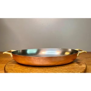 May include: A shallow, round copper pan with a polished metallic exterior and a dark, smooth interior surface. The pan features two curved, gold-toned metal handles attached to opposite sides, providing a sturdy grip for lifting.