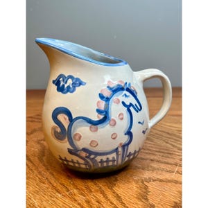 May include: A cream-colored ceramic pitcher featuring a hand-painted blue and pink horse design. The horse is depicted standing behind a fence with a blue cloud above. The pitcher has a blue rim, a curved handle, and a wide pouring spout.