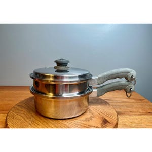 May include: Stainless steel double boiler pot set with a lid and two gray handles. The pot set is sitting on a wooden surface. The handles have a loop for hanging.