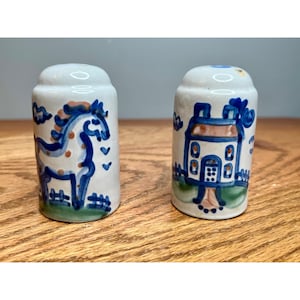 May include: A pair of cylindrical ceramic salt and pepper shakers with a white base. One shaker features a hand-painted blue horse with brown spots, while the other displays a blue house with green and brown accents, both set on a green ground.
