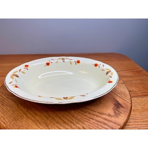 May include: Cream-colored oval serving bowl with a decorative rim. The rim features a pattern of red and yellow leaves and berries. The bowl has a silver-colored trim and sits on a wooden surface.
