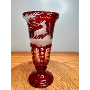 May include: A ruby red glass vase with a white etched design of a deer in a forest scene. The vase has a flared top, a cylindrical body with cut-out details, and a round base. The vase is approximately 8 inches tall.