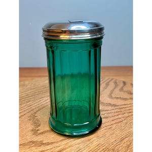 May include: A vintage green glass sugar dispenser with a ribbed design and a shiny silver metal lid. The cylindrical container is made of a vibrant green glass, adding a pop of color. The lid has a small opening for dispensing.