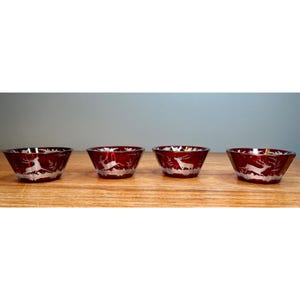 May include: A set of four small, circular, deep red glass bowls. Each bowl features a clear, etched design of a deer standing in a forest landscape. The bowls have a smooth, polished finish and are arranged in a horizontal row on a flat surface.