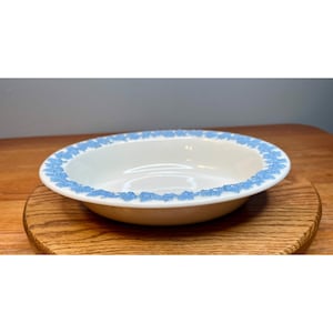 May include: A shallow, round white ceramic bowl featuring a decorative border of light blue floral patterns along the rim. The bowl has a smooth, glossy finish and a wide, flat edge that frames the central basin, creating a clean, circular design.