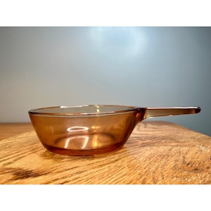 May include: A vintage amber-colored glass bowl with a handle. The bowl has a rounded shape and a smooth, transparent surface. The handle is an extension of the bowl, and the overall design is simple and functional. The bowl is likely for cooking or serving.