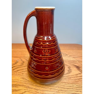 May include: A brown ceramic pitcher with a handle and a white rim. The pitcher has a bulbous body with horizontal bands and floral and dot designs. The pitcher is approximately 9 inches tall.