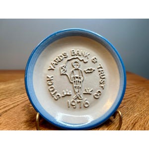 May include: A small, round, ceramic dish with a light blue rim and a beige center. The center features embossed text that reads "STOCK YARDS BANK & TRUST CO 1976" around a central figure holding a kite.