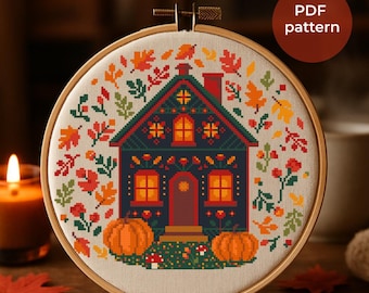 May include: A cross-stitch pattern of a house with orange pumpkins, surrounded by fall leaves in a wooden embroidery hoop. The house is dark blue with orange windows and a red door. The text "PDF pattern" is in a brown circle.