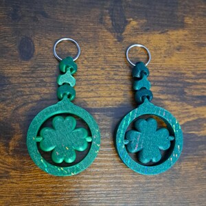 May include: Two green clover keychains with silver key rings. Each keychain features a four-leaf clover design within a circular frame. The keychains are made of a speckled green material, with a decorative top.