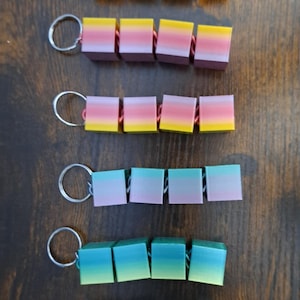 May include: Keychains with colorful, gradient-colored cube beads. Each keychain has a silver ring and a series of connected cubes in shades of pink, yellow, green, and blue. The keychains are arranged on a dark wooden surface.