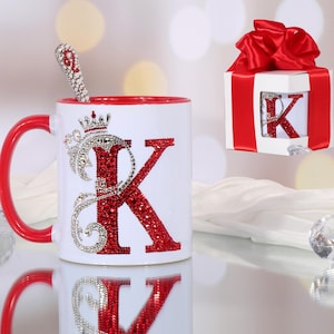 May include: White ceramic mug with a red interior and handle, adorned with a sparkling red letter "K" and a crown design. A matching spoon rests inside. A gift box with a red bow and a "K" is also present.