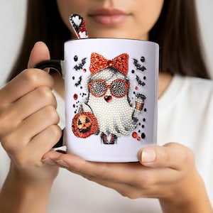 May include: White ceramic mug with a black handle and rim, featuring a rhinestone-studded ghost design. The ghost wears a red bow, sunglasses, and holds a pumpkin and a drink. A matching spoon rests inside.