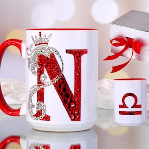 Può includere: Tazza in ceramica bianca con interno e manico rossi, con una grande lettera "N" rossa impreziosita da cristalli rossi e una corona argentata. Sono presenti anche una tazza più piccola con un simbolo della Bilancia e una confezione regalo con un fiocco rosso.