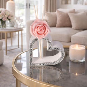 Könnte beinhalten: Ein weißes herzförmiges Dekorationsobjekt mit silbernem Strassbesatz, das eine rosa Rose und Holzstäbchen enthält. Eine Glas-Kerze steht daneben auf einem Glastisch. Wohnkultur.