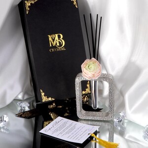 Peut inclure: Un coffret noir avec des détails dorés et le logo "MB Crystal" est à côté d'un vase décoratif argenté contenant une fleur et des roseaux. Une carte avec un pompon jaune est également présente, suggérant un article de luxe.