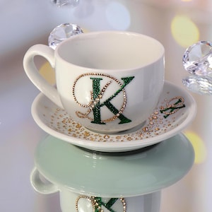 Op de afbeelding: Witte theekop en schotel set met een groen en goud letter "K" ontwerp. De theekop en schotel zijn versierd met gouden en heldere strass steentjes. De set staat op een reflecterend oppervlak met decoratieve kristallen.