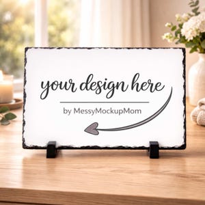 May include: A rectangular white slate sign with the text "your design here" in a script font, and the text "by MessyMockupMom" below. The sign is supported by two black stands and sits on a wooden surface.