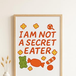 May include: A framed print with the text "I AM NOT A SECRET EATER" in orange. The print features illustrations of candy, including a green gummy bear, a wrapped orange candy, a lollipop, and a swirl.