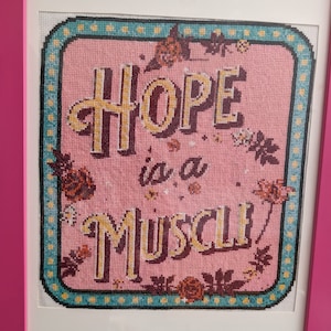 May include: A framed cross-stitch with a pink background and a teal border. The cross-stitch reads "Hope is a Muscle" in a stylized font with floral accents.