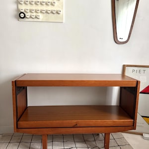 May include: A rectangular wooden side table with a warm brown finish, featuring two open shelves and four small wheels attached to the base. The table has a clean, simple construction with visible joinery details on the side panels.