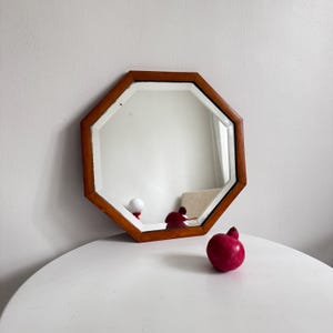 Könnte beinhalten: Ein achteckiger Spiegel mit einem braunen Holzrahmen. Der Spiegel steht auf einem weißen Tisch, mit einem roten dekorativen Granatapfel und anderen kleinen Objekten. Der Spiegel reflektiert die Objekte auf dem Tisch.