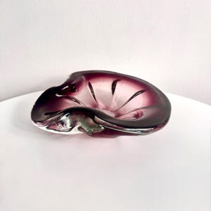 Könnte beinhalten: Ein Vintage-Aschenbecher aus mundgeblasenem Glas in tiefem Lila. Der Aschenbecher hat eine einzigartige, organische Form mit einem ausgestellten Rand und einer glatten, glänzenden Oberfläche. Der Innenraum ist in einem helleren, durchscheinenden Lila gehalten.
