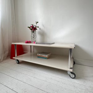 May include: A cream-coloured, two-tiered rolling cart with chrome legs and wheels. The top shelf holds a laptop, a vase of pink flowers, and a stack of books. A red stool is visible on the left.
