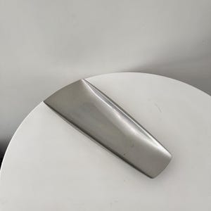 May include: A long, rectangular silver-toned metal tray with a smooth, polished finish. The tray features slightly curved edges and a shallow, concave centre, presenting a sleek, uniform appearance with a metallic sheen throughout its surface.