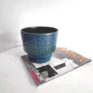 Op de afbeelding: Een blauwgroene keramische plantenbak met een getextureerde, gedruppelde glazuur. De plantenbak heeft een donker interieur en een getrapte basis. Hij ligt op een tijdschrift met de tekst "CASA VOGUE" en "HOME STORIES n.36".