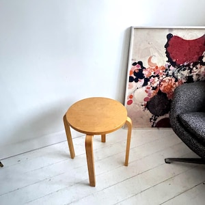 May include: A round, light-colored wooden stool featuring three curved legs that extend from the underside of the circular seat. The stool has a smooth, natural wood grain finish and a simple, functional design with a flat top surface.
