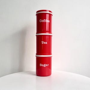 May include: Three red kitchen canisters stacked on top of each other, labelled "Coffee", "Tea", and "Sugar" in white text. Each canister has a white band around the top. The canisters are on a white surface.