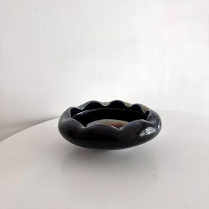 Könnte beinhalten: Ein dunkler, glänzender, ovaler Aschenbecher mit gewelltem Rand. Der Aschenbecher hat eine tiefschwarze Farbe mit schillernden Farbtönen. Das Innere ist glatt und reflektierend, auf einer weißen Oberfläche.