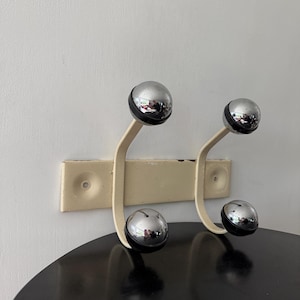 May include: A wall-mounted coat rack featuring a rectangular cream-coloured metal base with two curved arms. Each arm supports two spherical hooks, totalling four hooks. The hooks are chrome-finished spheres with black circular accents at the base.